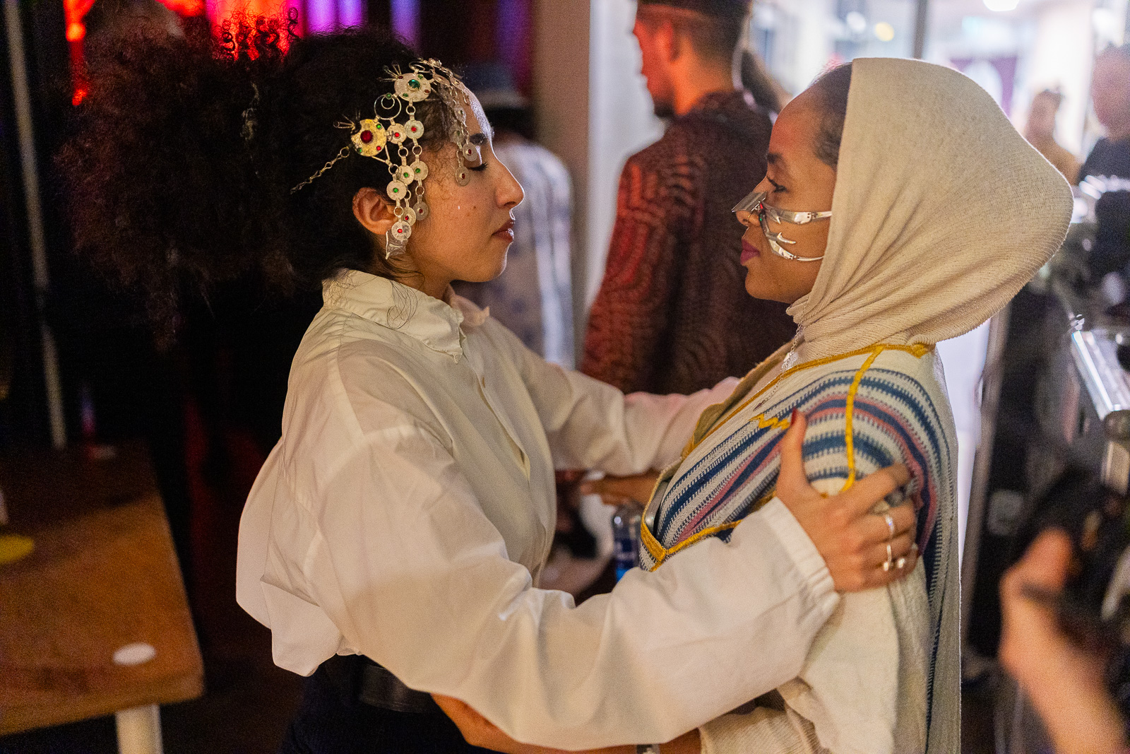 This photo shows two people holding each other. On the left you see a woman with her eyes closed holding the shoulders of the other woman. She is wearing an ornament on her head. On the right, you see a woman with a headscarf and an ornament on her nose. She is looking at the women on the left and holder her arms.